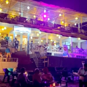 A lively nighttime scene at Anjuna Beach, Goa. The image features a two-story establishment with colorful lights on the upper deck, where people are standing and sitting. The lower level appears to be a restaurant or bar with patrons seated at tables. In the foreground, more people are gathered on the beach, some standing and others seated around candle-lit tables. The dark sky and ambient lighting create a vibrant, social atmosphere by the sea.