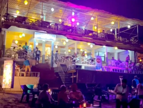 A lively nighttime scene at Anjuna Beach, Goa. The image features a two-story establishment with colorful lights on the upper deck, where people are standing and sitting. The lower level appears to be a restaurant or bar with patrons seated at tables. In the foreground, more people are gathered on the beach, some standing and others seated around candle-lit tables. The dark sky and ambient lighting create a vibrant, social atmosphere by the sea.