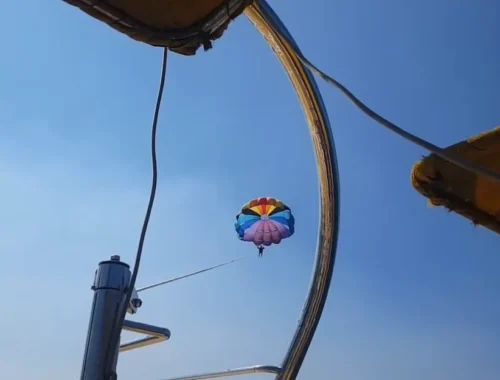 Parasailing adventure at Anjuna Beach, Goa, with a colorful parachute soaring in the clear blue sky, viewed from under the shade of a beach umbrella.