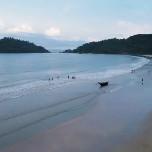 Panoramic view of Palolem Beach in Goa, featuring a gently curving shoreline with light-colored sand meeting the calm blue waters of the Arabian Sea. A small island is visible in the distance, and several people are scattered along the shore, enjoying leisure activities. A traditional boat is anchored on the shore in the foreground under an overcast sky.