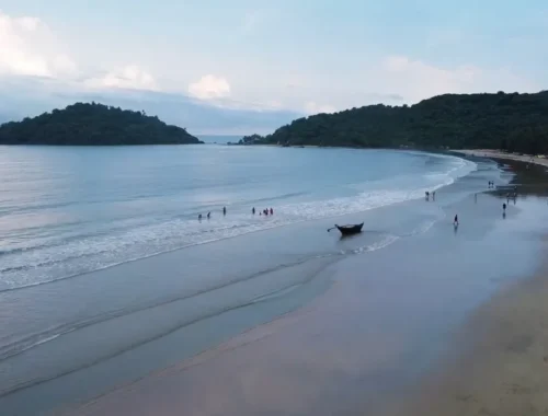Panoramic view of Palolem Beach in Goa, featuring a gently curving shoreline with light-colored sand meeting the calm blue waters of the Arabian Sea. A small island is visible in the distance, and several people are scattered along the shore, enjoying leisure activities. A traditional boat is anchored on the shore in the foreground under an overcast sky.